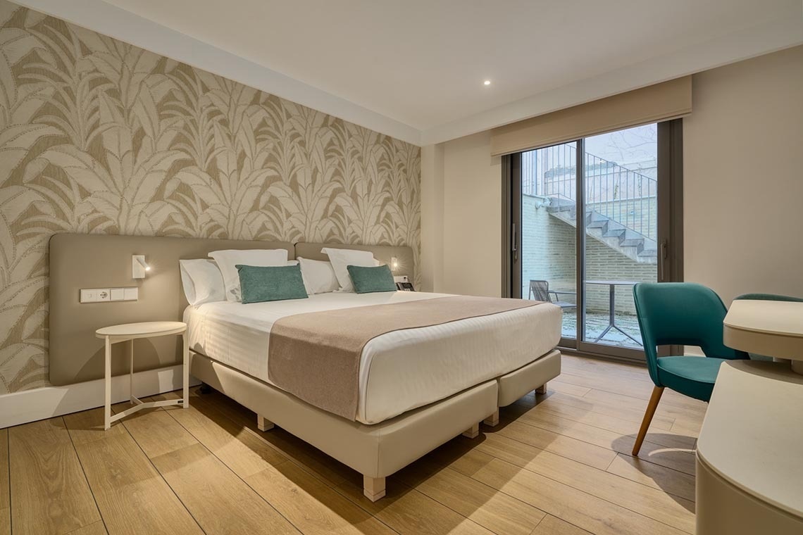 Una habitación moderna con una cama doble, papel tapiz decorado con hojas, y una puerta de cristal que da a un patio con escaleras.