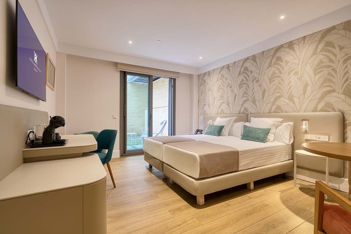 Luminosa habitación de hotel con dos camas, escritorio, televisor y puerta de cristal que da al exterior, decorada con papel pintado de hojas.