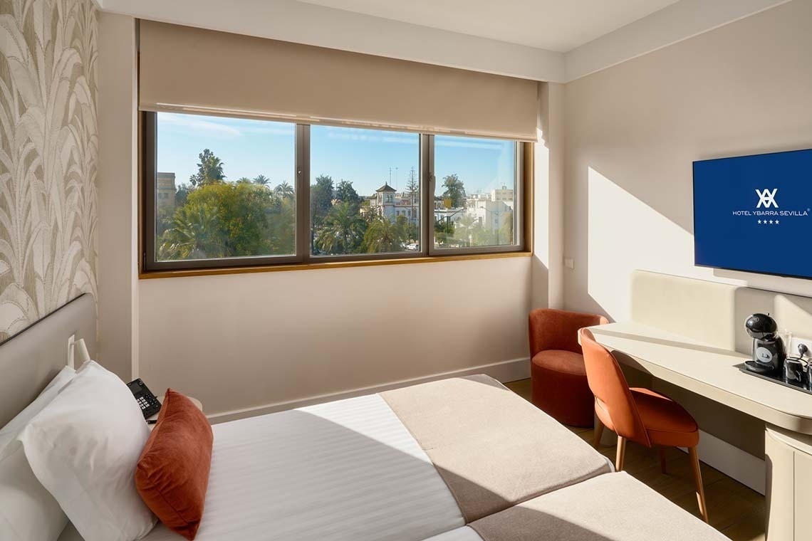 La imagen muestra una luminosa habitación de hotel con una cama, escritorio, televisión y una gran ventana que ofrece vistas a un paisaje urbano arbolado.