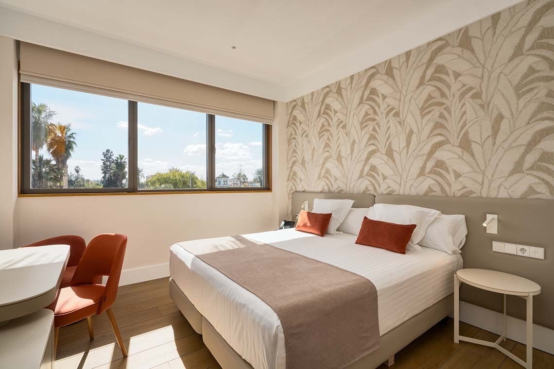 Un luminoso dormitorio con una cama hecha, papel pintado de hojas botánicas y una gran ventana con vistas a palmeras y vegetación.