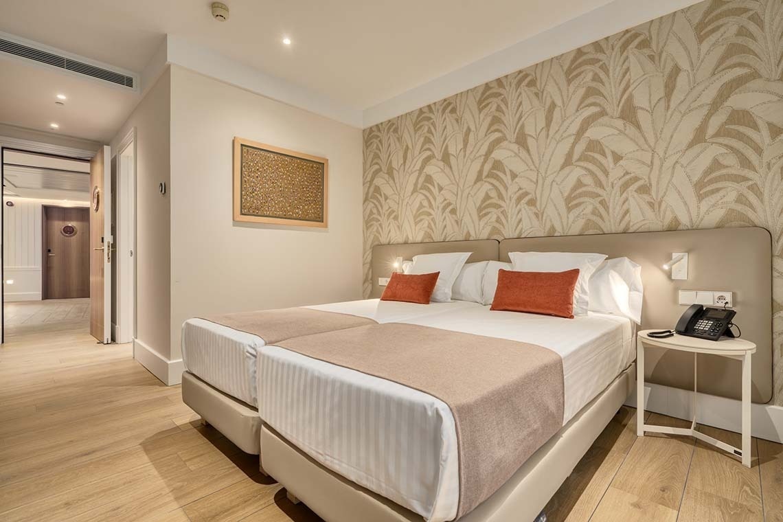 Una habitación de hotel con dos camas individuales, una pared decorada con papel pintado de hojas, suelo de madera y un pasillo visible a la izquierda.