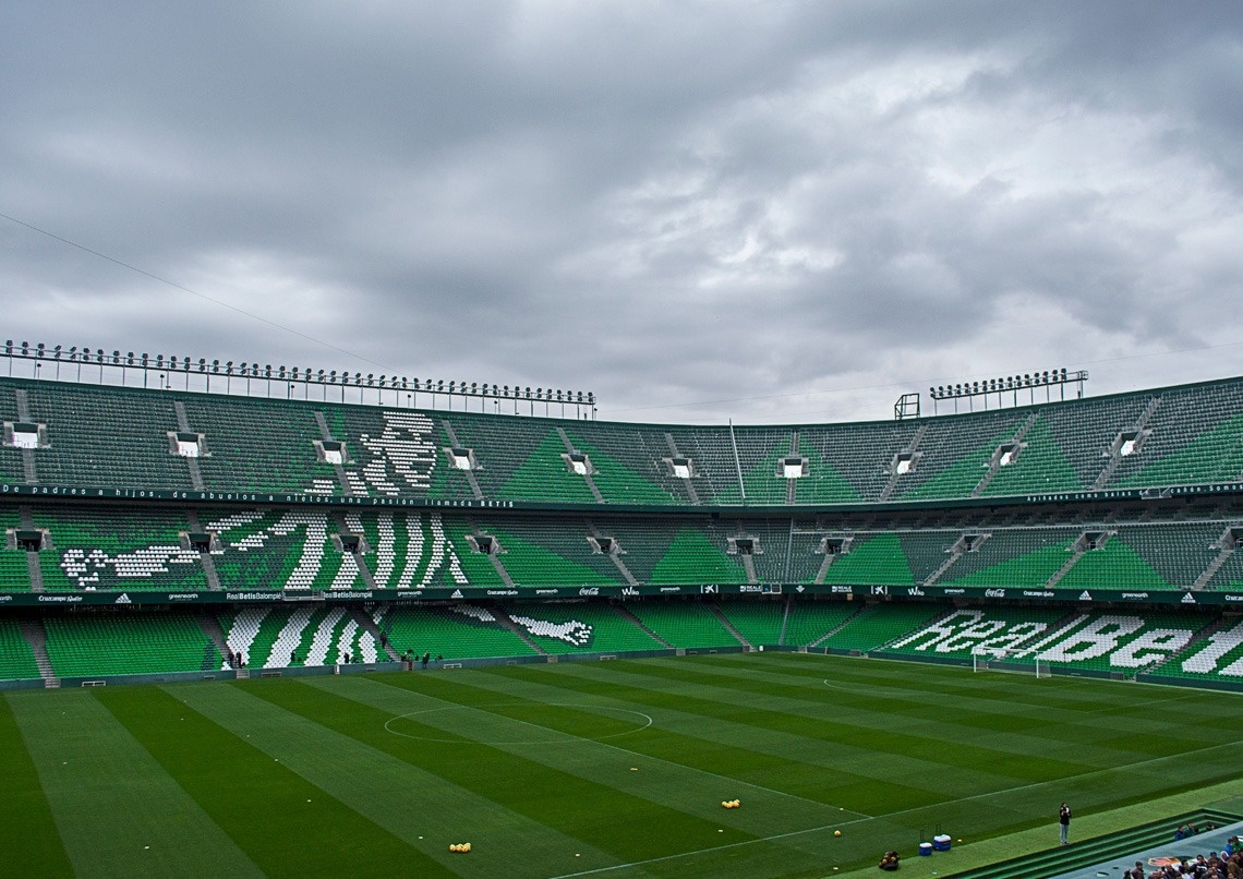 Un estadio de fútbol vacío, con sus gradas verdes y blancas y césped bien cuidado, se extiende bajo un cielo nublado.