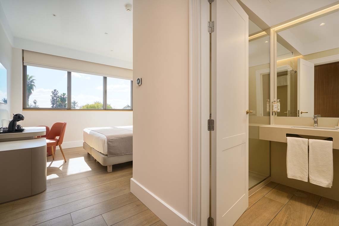 Una luminosa y moderna habitación de hotel con cama, escritorio y un baño visible, destacando grandes ventanales con palmeras en el exterior.