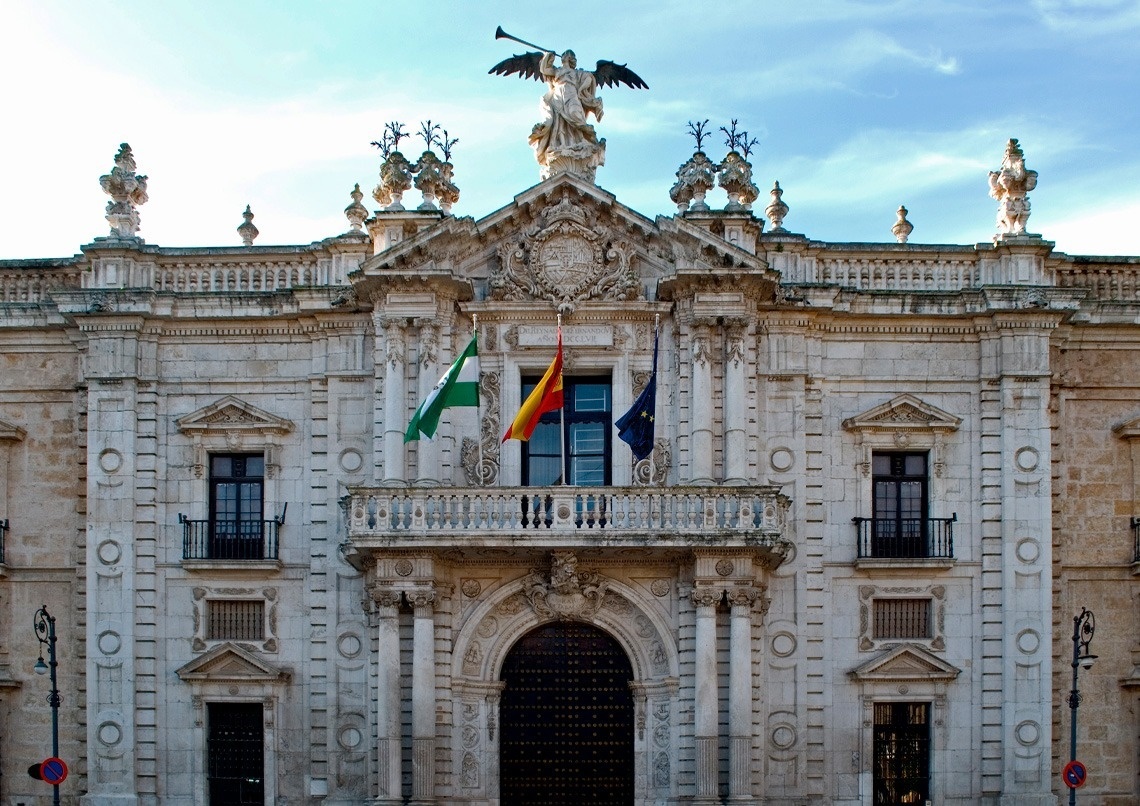 Este es un primer plano de la fachada ornamentada del Ayuntamiento de Sevilla, adornada con tres banderas (Andalucía, España y la Unión Europea) y una estatua en la parte superior, bajo un cielo parcialmente nublado.