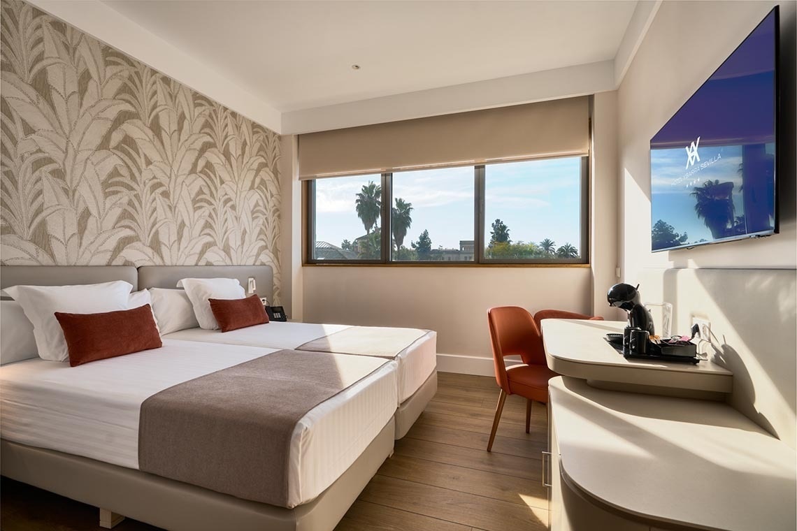 Una moderna habitación de hotel cuenta con dos camas individuales, un ventanal con vistas a palmeras, un televisor y una cafetera, realzada por una pared con papel pintado de hojas.