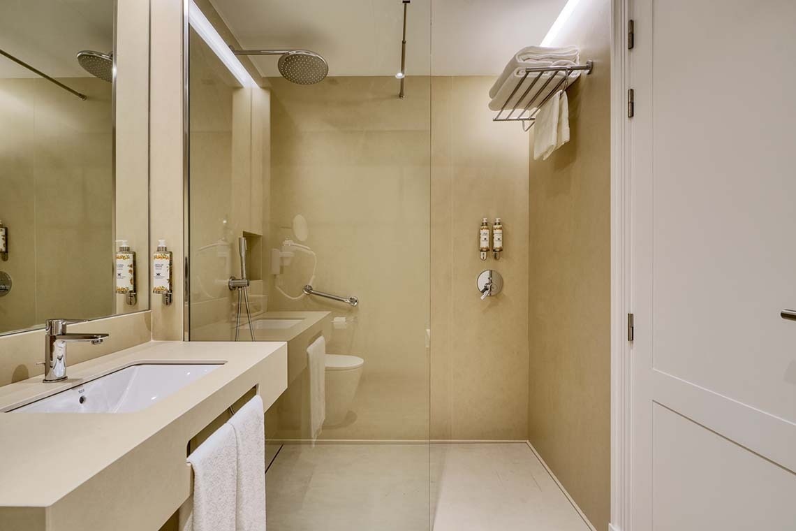 Un moderno y funcional cuarto de baño en tonos crema, con lavabo, espejo, una ducha accesible con mampara de cristal y un inodoro.