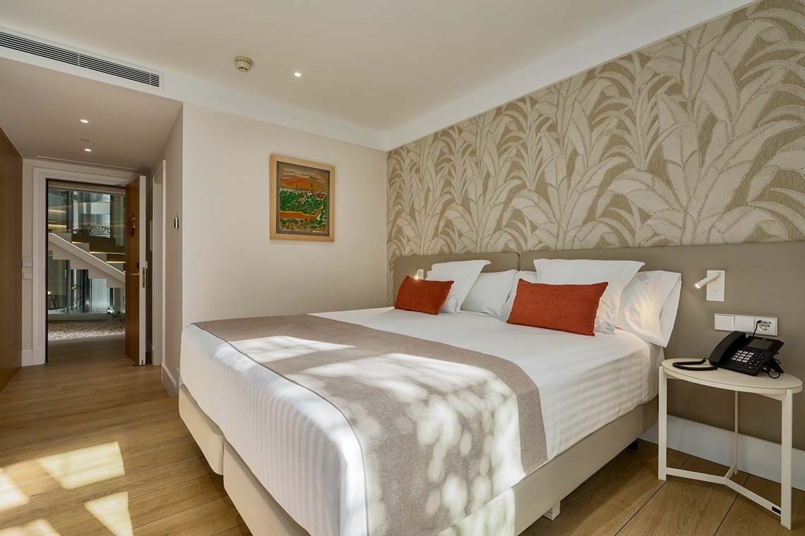 Una habitación de hotel moderna y luminosa con una cama grande, papel tapiz estampado y un pasillo visible con escaleras.