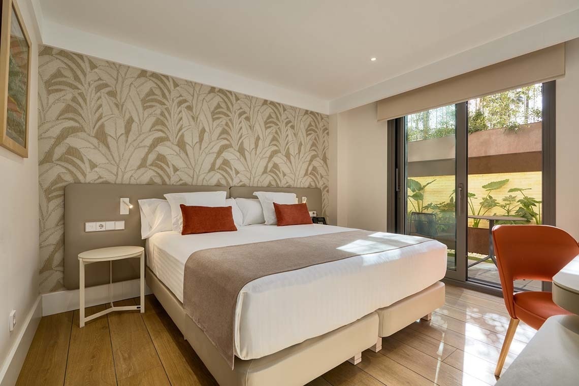Una moderna habitación de hotel cuenta con una cama grande, una pared decorada con papel pintado de hojas, y una puerta de cristal que da a un patio exterior con vegetación.