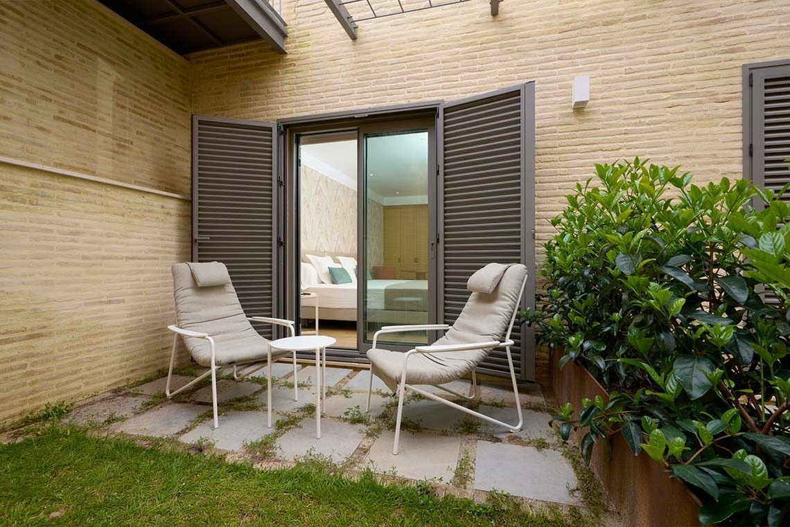 Patio exterior con dos sillones y una mesa pequeña, rodeado de vegetación y una pared de ladrillo, con acceso a un dormitorio a través de una puerta de vidrio.