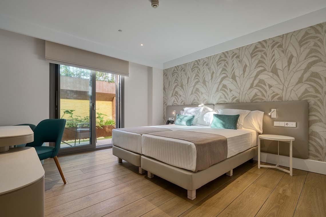 Una moderna habitación de hotel con dos camas individuales, una pared decorada con papel pintado de hojas y una puerta corredera de cristal que da a un patio exterior con vegetación.