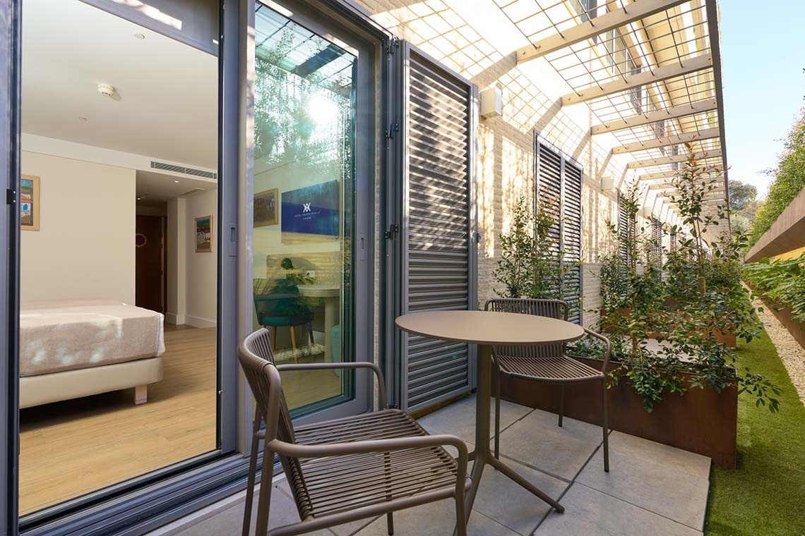 Una habitación de hotel se abre a un patio exterior amueblado con una mesa y sillas, rodeado de plantas en jardineras y cubierto parcialmente por una pérgola.