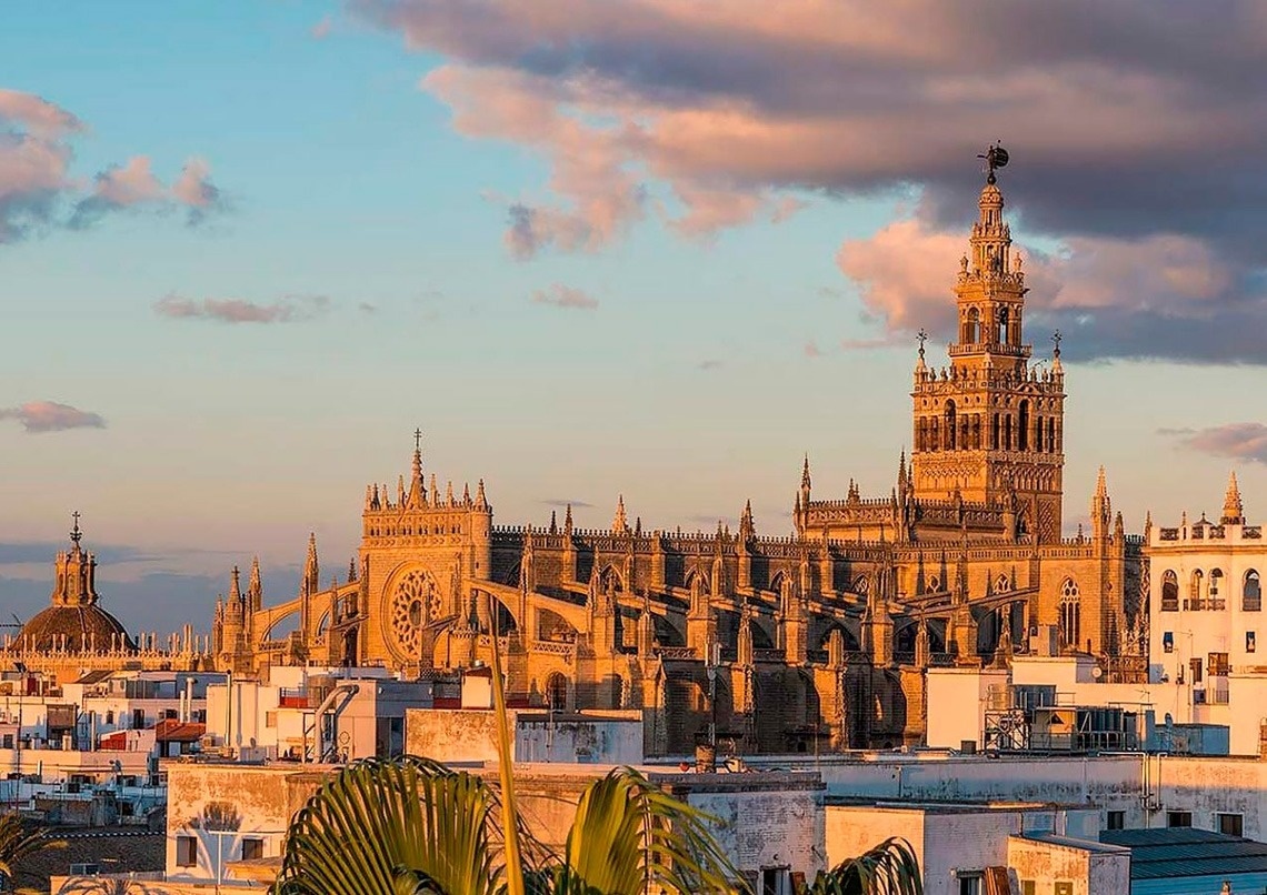 Una majestuosa catedral gótica se alza imponente bajo la luz dorada del atardecer, con su campanario dominando un cielo de suaves nubes rosadas y azules.
