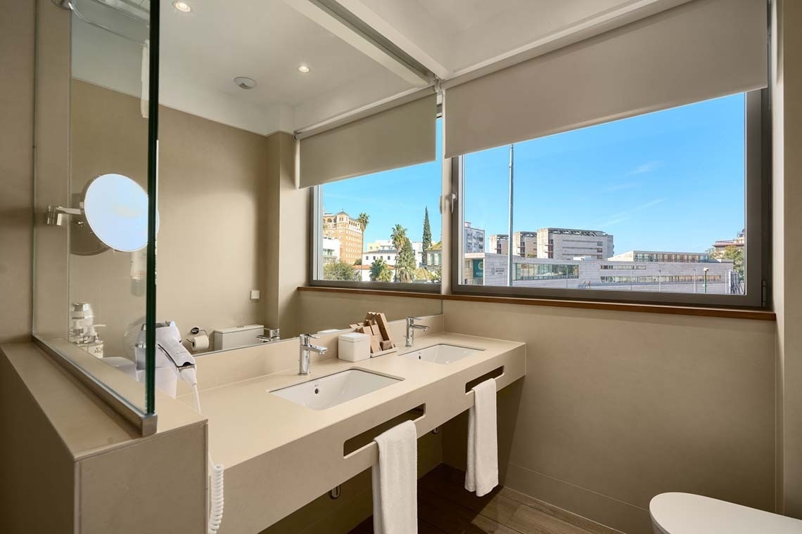 Un baño moderno con lavabo doble presenta una gran ventana con persianas enrollables que ofrece vistas a edificios y un cielo azul claro.