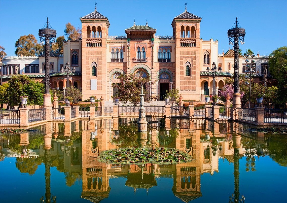Un pabellón de estilo mudéjar con torres se refleja perfectamente en un estanque tranquilo bajo un cielo azul.