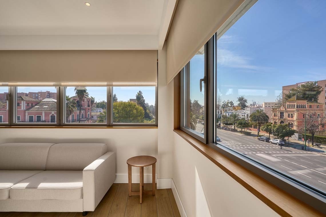 Un interior luminoso con amplios ventanales que ofrecen vistas a una soleada calle urbana con edificios y árboles.