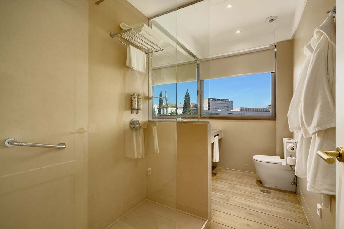 Un baño moderno y luminoso con ducha de cristal, inodoro, suelo de madera y una ventana con vista al exterior.