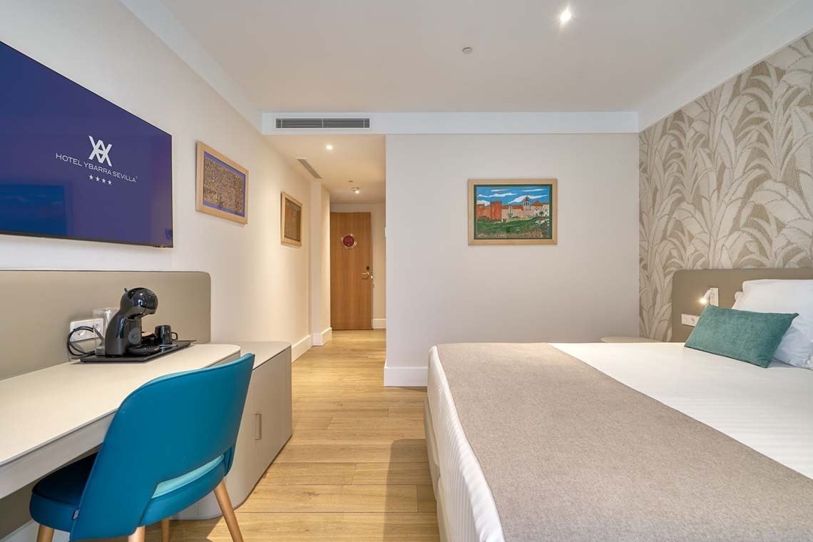 Una habitación de hotel moderna y luminosa con cama, escritorio con cafetera y televisor, y un pasillo que se extiende al fondo.