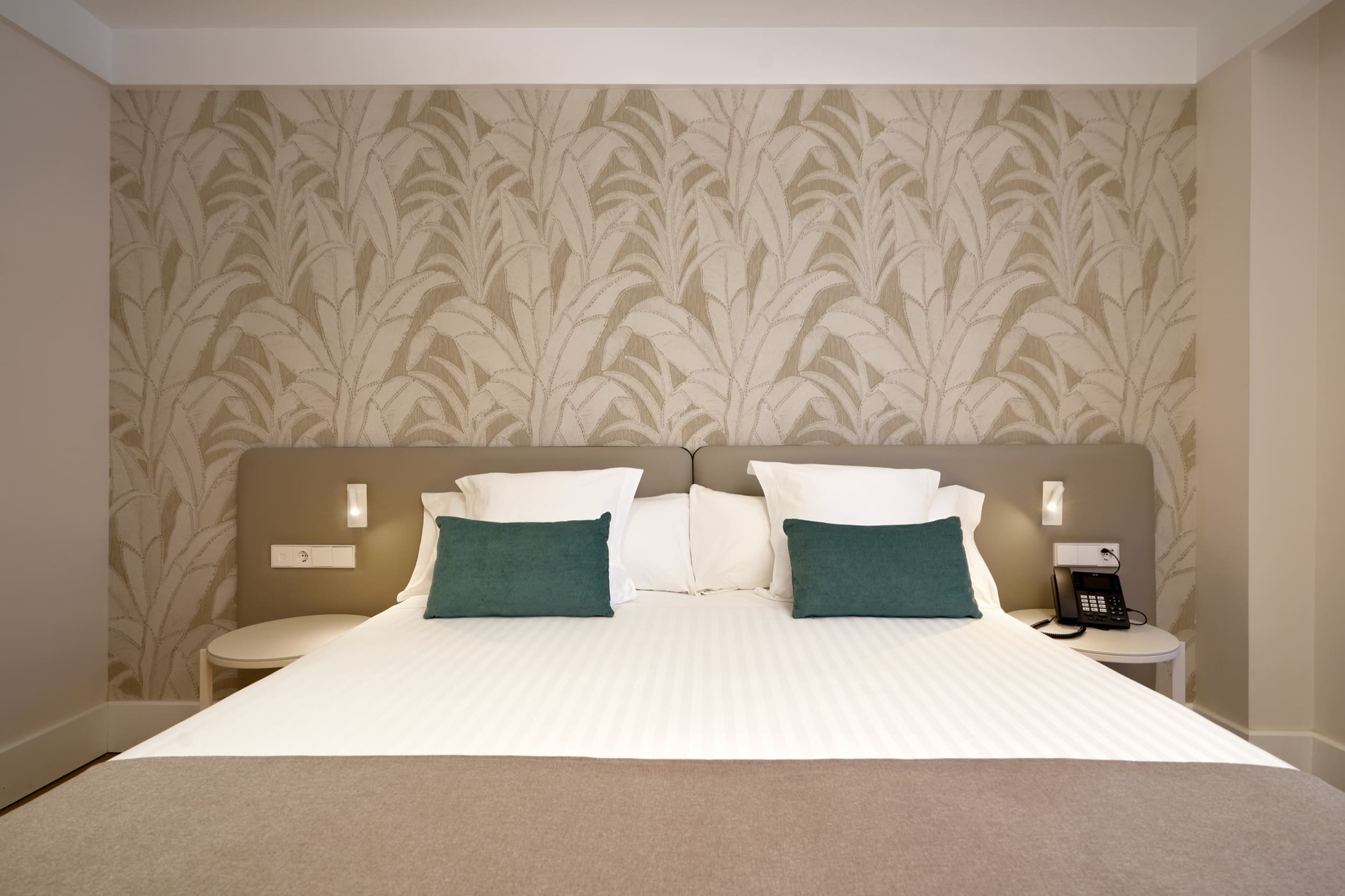 Comfortable bed with green accent pillows at Ybarra Sevilla, featuring botanical wallpaper and bedside table with phone.