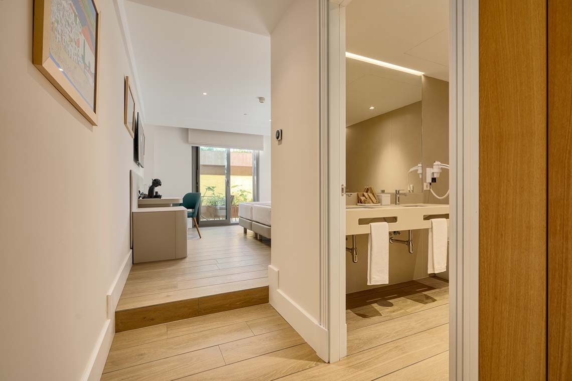 Un pasillo con suelo de madera clara conecta una moderna habitación con balcón y un baño con doble lavabo.