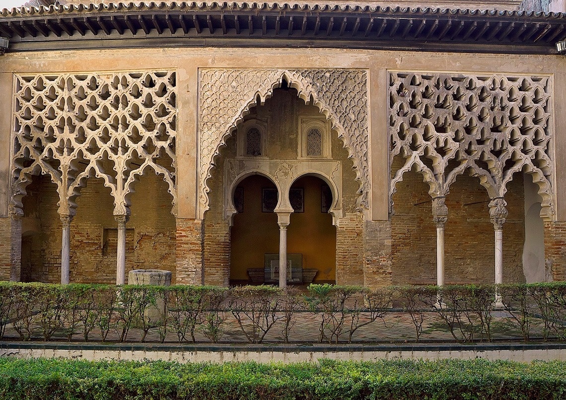 Una galería arquitectónica de estilo mudéjar, ricamente decorada con arcos intrincados, celosías y columnas, enmarca un patio con setos cuidadosamente podados.