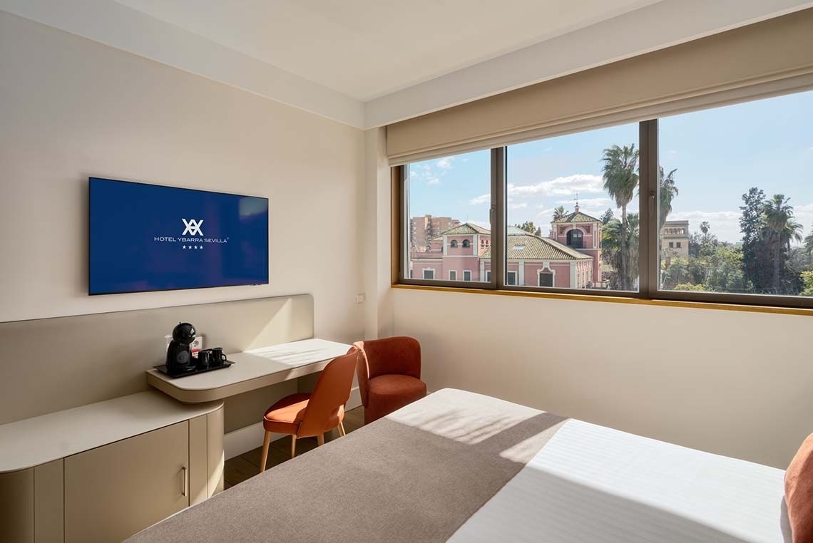 Una moderna y luminosa habitación de hotel cuenta con un televisor, una zona de escritorio con cafetera y una gran ventana que ofrece vistas a edificios históricos y palmeras.