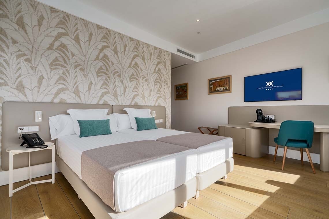 Una moderna y luminosa habitación de hotel presenta dos camas individuales, una pared con papel pintado de hojas y un escritorio con televisor y una silla turquesa.