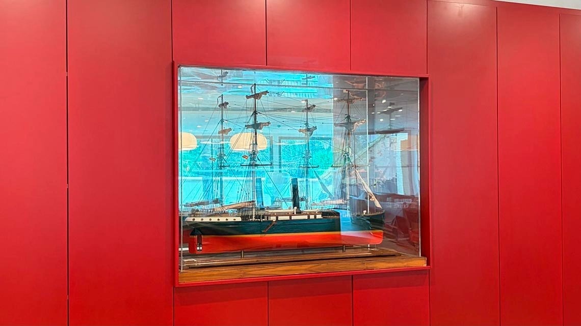 Una maqueta de un barco de vela con chimenea se exhibe en una vitrina de cristal incrustada en una pared roja brillante, con reflejos visibles en el cristal.