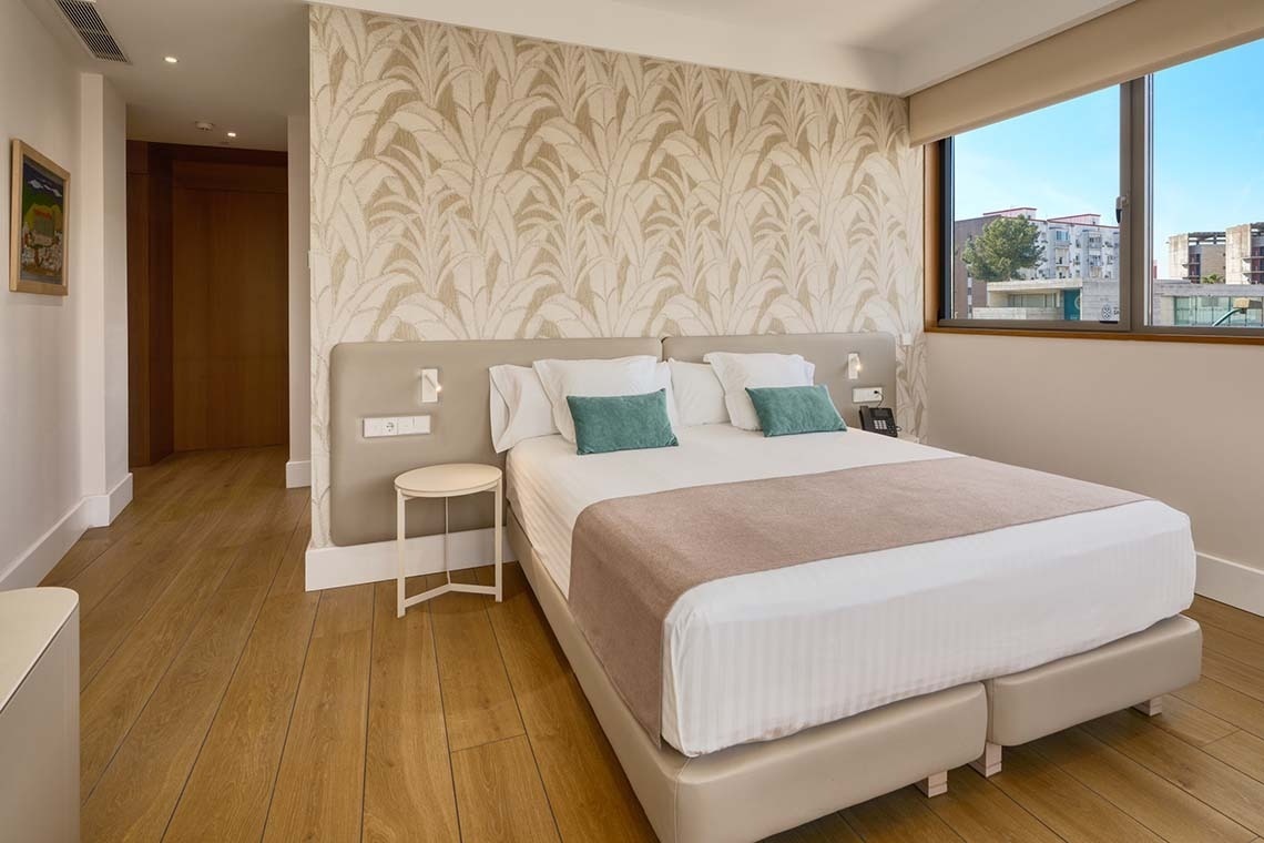 Una habitación de hotel moderna y luminosa presenta una cama grande con cabecero claro, papel tapiz con patrón de hojas, suelo de madera y un gran ventanal con vista a edificios y vegetación urbana.