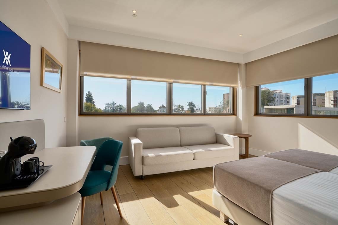 Una luminosa habitación con grandes ventanales que ofrecen una vista exterior, un sofá, una mesa con cafetera y una cama.