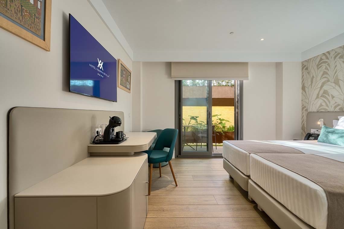 La imagen muestra una habitación de hotel moderna y luminosa con camas gemelas, un escritorio con cafetera, televisor y acceso a un balcón con plantas.