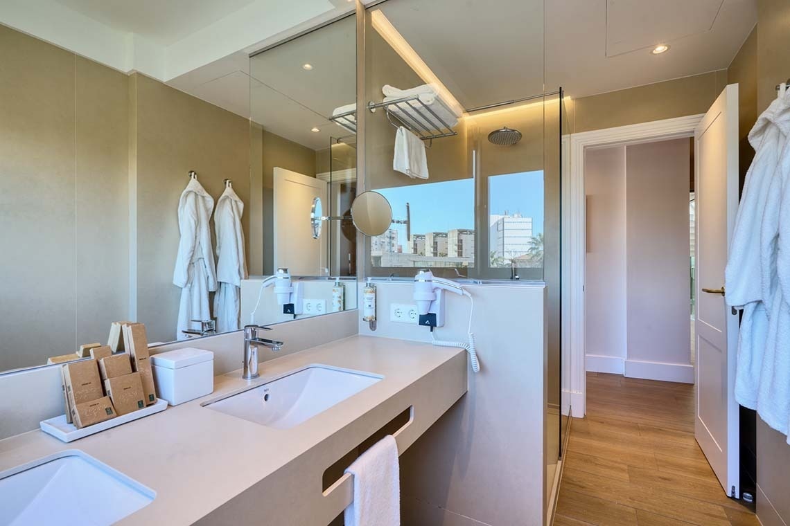 Un baño moderno con doble lavabo, un gran espejo, una ducha acristalada y albornoces colgados, ofreciendo una vista exterior a través de la ventana de la ducha.