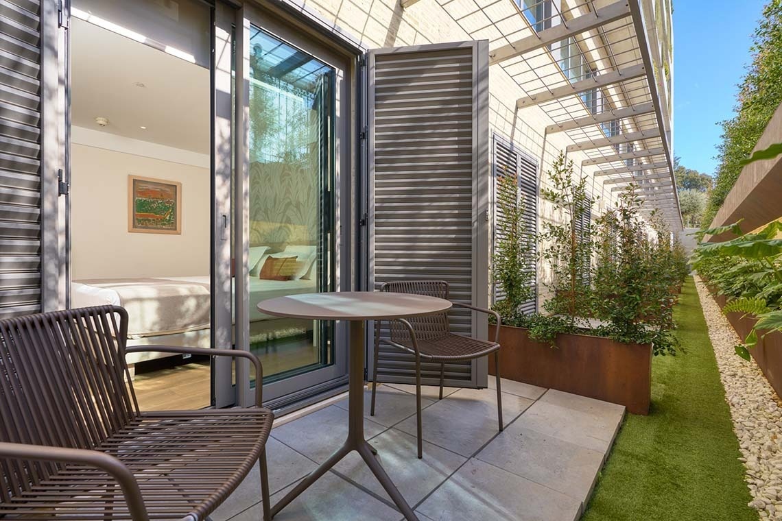 Una terraza exterior moderna con mesa y sillas, conectada a una habitación luminosa a través de una puerta de cristal, se extiende hacia un estrecho jardín con plantas y césped artificial bajo una pérgola.