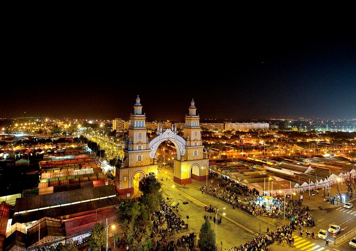 Una imponente portada ricamente iluminada preside una vibrante feria nocturna, repleta de gente y casetas, con las luces de la ciudad extendiéndose en la oscuridad.