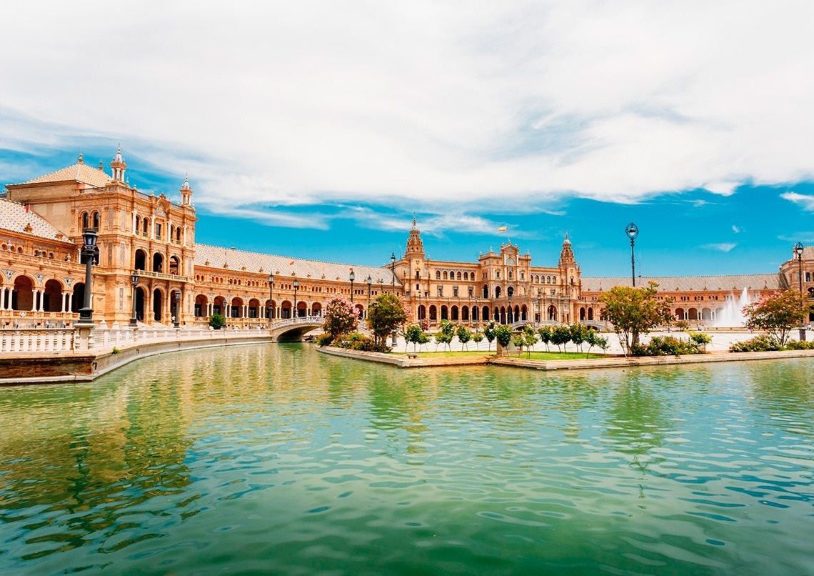 Un impresionante edificio histórico con múltiples arcos y torres se alza frente a un canal con un puente, todo bajo un cielo azul y nubes.