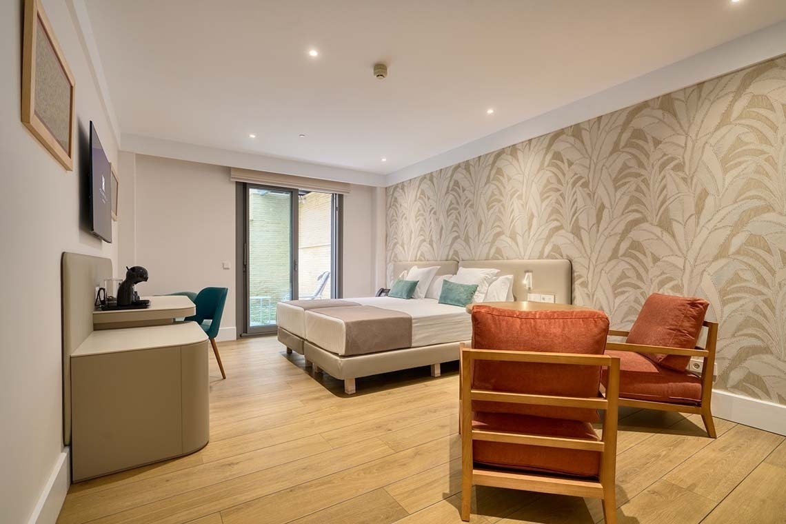 Una moderna habitación de hotel presenta dos camas individuales con cabecero claro, un escritorio con silla azul, dos sillones de madera con cojines naranjas y una pared decorada con papel tapiz de hojas claras, todo sobre un suelo de madera clara.