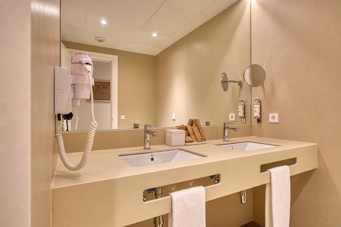 Un moderno cuarto de baño presenta un tocador doble de color beige con lavabos integrados, un espejo grande, un secador de pelo montado en la pared y toallas blancas limpias.