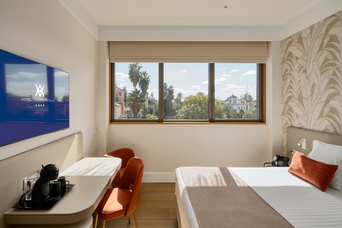 Un luminoso dormitorio cuenta con una cama grande vestida con cojines naranjas, una pared decorada con papel pintado de hojas, suelo de madera y una amplia ventana con vistas a palmeras.