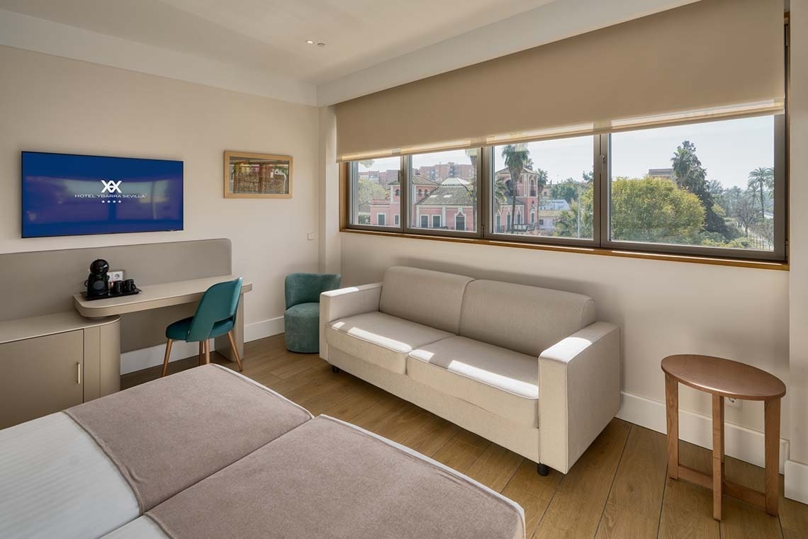 Una luminosa habitación de hotel cuenta con una cama, un área de trabajo con televisión, un sofá beige y una amplia ventana con vistas a edificios y árboles de la ciudad.