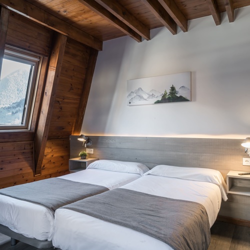 Acollidor dormitori amb dos llits individuals, parets i sostre de fusta inclinats, i una finestra amb vistes a una muntanya nevada.