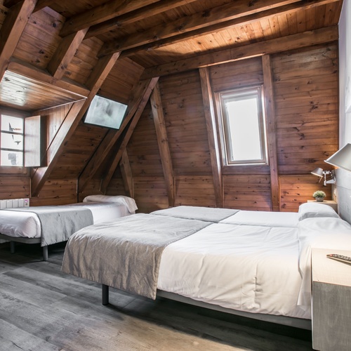 Una habitació de sostre abuhardillat amb revestiment de fusta, que conté un llit de matrimoni format per dos llits individuals i un llit individual més petit, amb finestres i un terra de parquet gris.