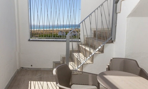 Ein heller Innenraum zeigt eine Treppe und ein vergittertes Fenster mit Meerblick auf eine Küstenlandschaft, ergänzt durch Sitzmöbel im Vordergrund.