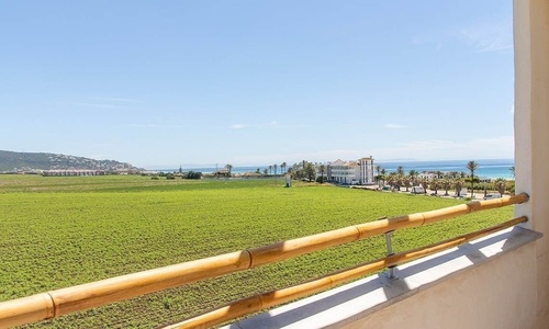 Ein Blick von einem Balkon zeigt ein weites grünes Feld, ein Gebäude mit Palmen am Meer und eine Hügellandschaft im Hintergrund unter blauem Himmel.