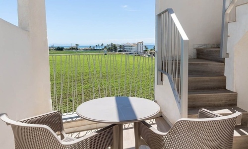 Ein Balkon mit Tisch und Stühlen bietet eine weite Aussicht auf eine grüne Wiese, das Meer und Palmen, wobei rechts eine Treppe sichtbar ist.