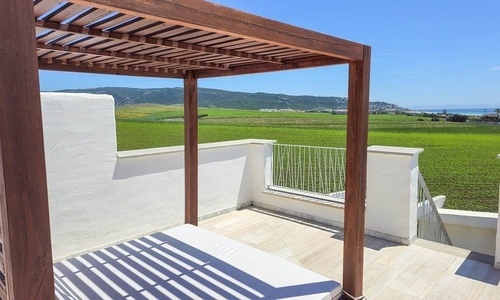 Eine sonnige Terrasse mit Holzpergola bietet einen weiten Blick über grüne Felder, ein entferntes Meer und Hügel.