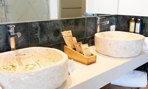 Ein Badezimmer zeigt einen Doppelwaschtisch mit zwei hellen, steinernen Waschbecken, modernen Chromarmaturen und Natur-Pflegeprodukten vor einer dunkelgrünen Marmorwand.