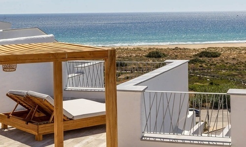 Eine weiße Terrasse mit hölzerner Pergola und Sonnenliegen bietet einen weiten Blick auf einen Sandstrand und den blauen Ozean.