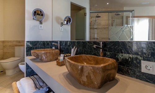 Ein modernes Badezimmer präsentiert sich mit zwei steinernen Waschbecken auf einem Waschtisch, einem großen Spiegel, einer Dusche mit Glastür, einer Toilette und einem Bidet.