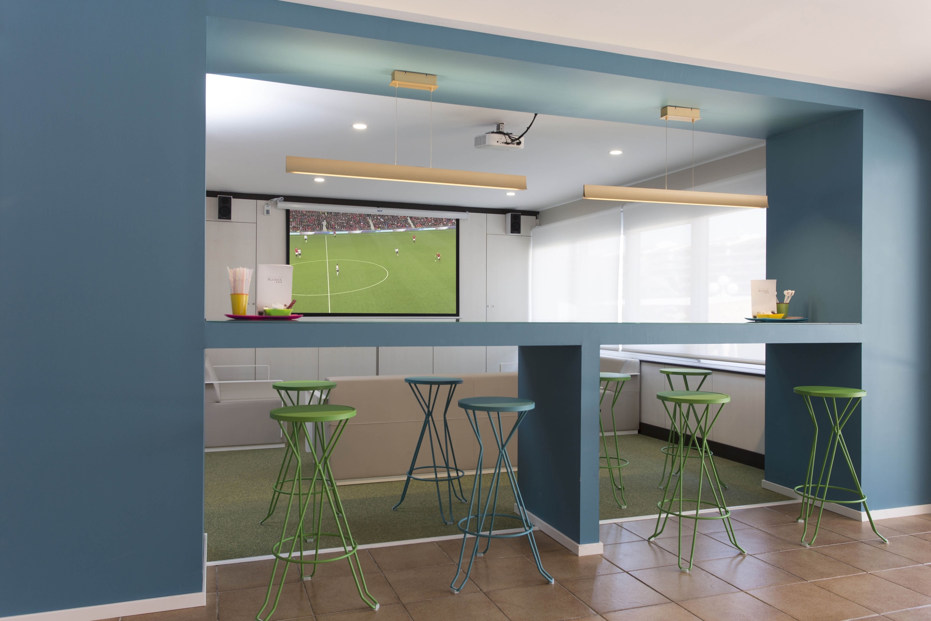 un bar con taburetes verdes y una pantalla de fútbol en la pared