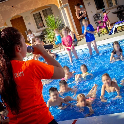 una mujer con una camiseta naranja con la palabra equipo en la espalda habla a un grupo de niños en una piscina