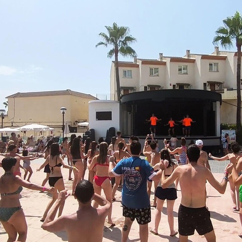 un grupo de jóvenes bailan en una piscina mientras un hombre con una camisa azul con la palabra ibiza en la espalda